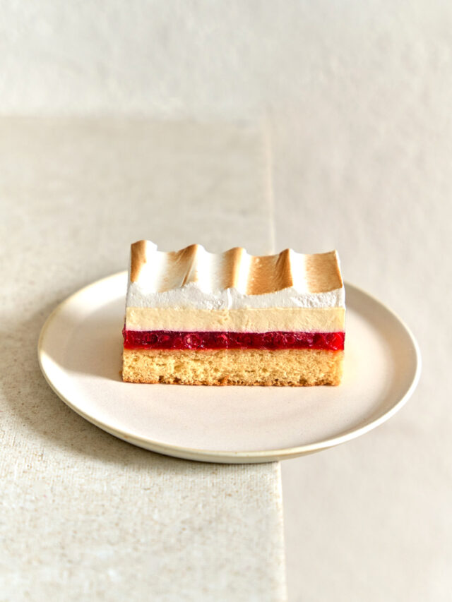 Eine geschichtet Schnitte mit hellem Biskuit, Ribiseln, Vanillecreme und Eischauem liegt auf einem Teller.