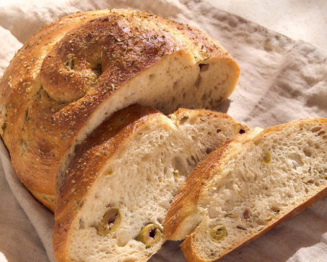 Ein helles Brot mit grünen Oliven wurde in Scheiben geschnitten. Alles liegt auf einem beigen Leinentuch.