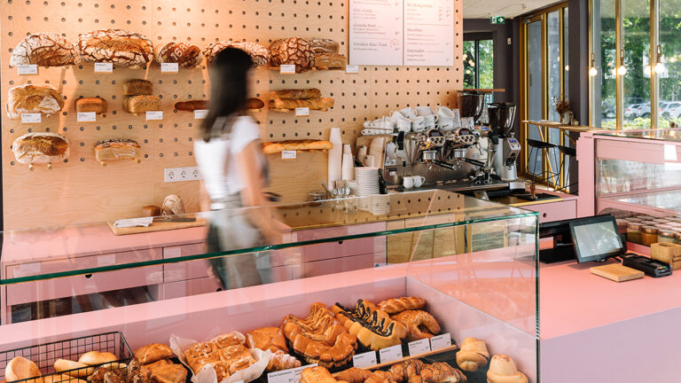 Ein frontal fotografiertes Brotregal. Am Rand sieht man verschwommen eine Mitarbeiterin der Bäckerei Sorger sowie eine Kaffeemaschine.