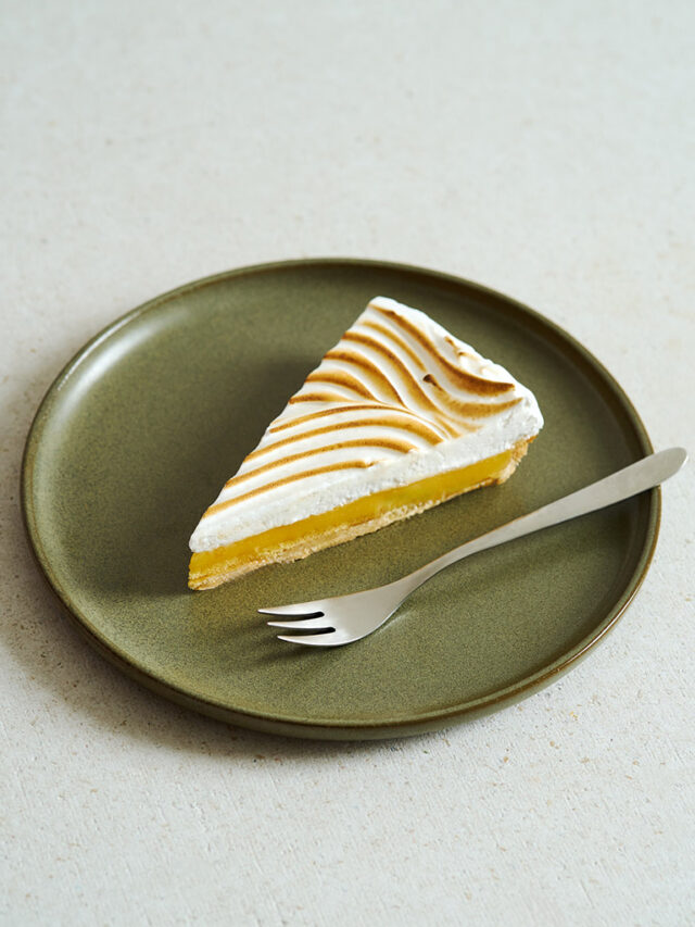 Ein Stück Tarte mit Mürbeteig, Zitronencreme und flambiertem Eischnell liegt mit einer Gabel auf einem grünen Teller.
