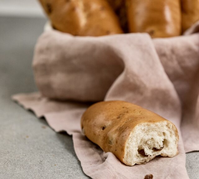 Mehrere Rosinenweckerl liegen im Hintergrund in einem Korb, der mit einem rosa Tuch ausgelegt ist. Im Vordergrund liegt ein angerissenes Rosinenweckerl.