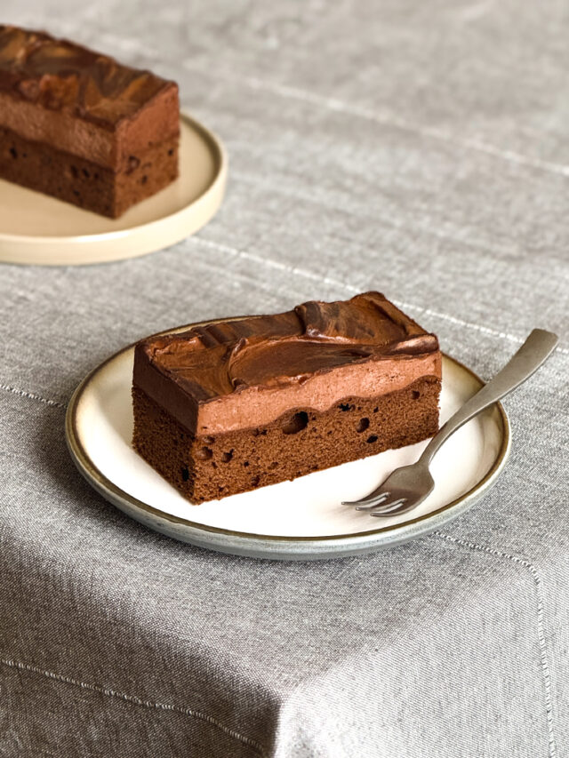 Ein Stück Schnitte aus Schokobiskuit mit Schokoladenmousse liegt auf einem weißen Teller. Im Hintergrund sieht man ein zweites Stück.