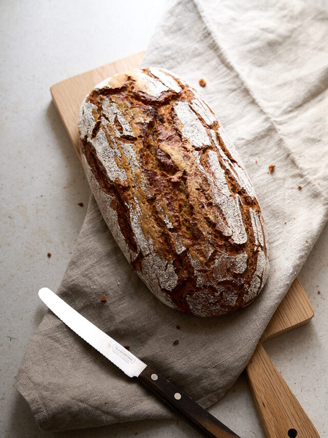 Ein kleines Schwarzbrot liegt zusammen mit einem Leinentuch auf einem Holzbrett. Daneben liegt ein Brotmesser.