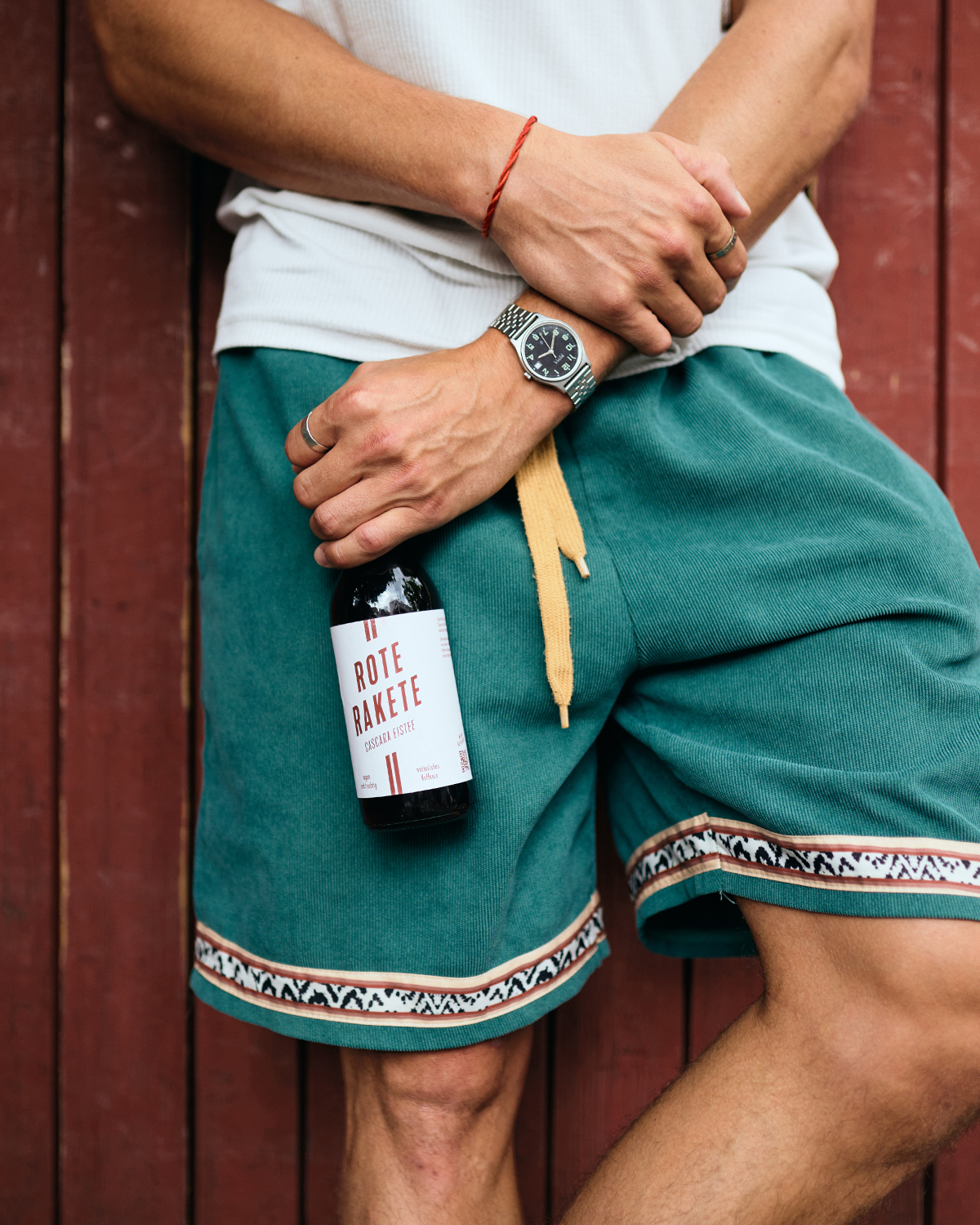 Mann mit grüner Hose lehnt an roter Wand und hält eine Flasche Rote Rakete.