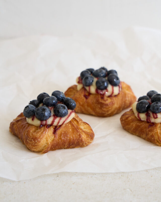 Drei Plunderecken mit Vanillecreme und Heidelbeeren liegen auf weißem Backpapier.